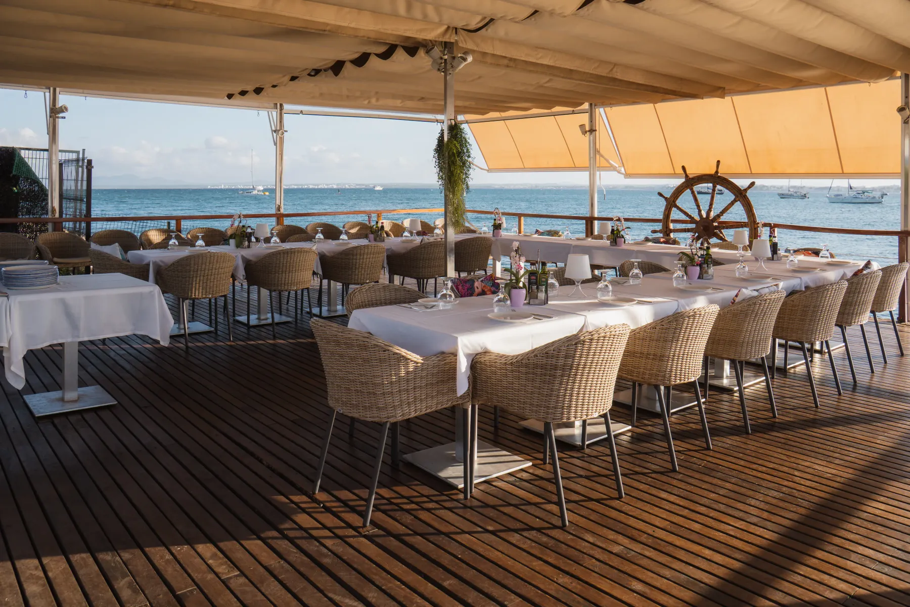Terraza de madera con mesas vestidas y timón de barco frente al mar